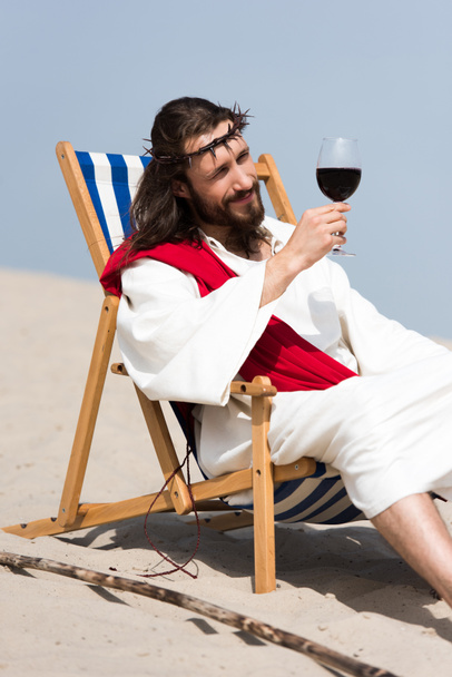 alegre Jesús en túnica y faja roja descansando sobre una tumbona y mirando un vaso de vino tinto en el desierto
 - Foto, imagen