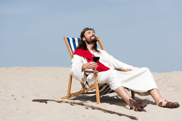 Иисус в халате и красной ленте отдыхает на шезлонге с бокалом красного вина в пустыне
 - Фото, изображение