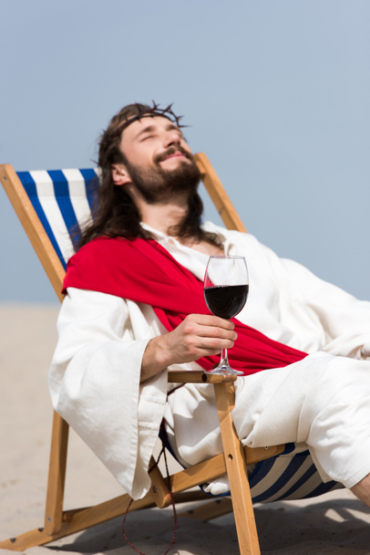 selective focus of cheerful Jesus in robe and red sash resting on sun lounger with glass of red wine in desert - 写真・画像