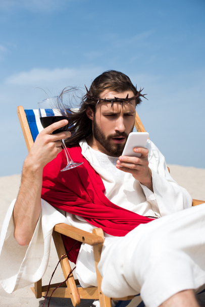 surprised Jesus resting on sun lounger with glass of wine and looking at smartphone in desert - Photo, Image