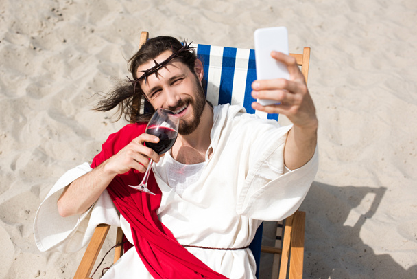 lächelnder Jesus, der sich auf Sonnenliegen ausruht, Wein trinkt und in der Wüste ein Selfie mit dem Smartphone macht - Foto, Bild