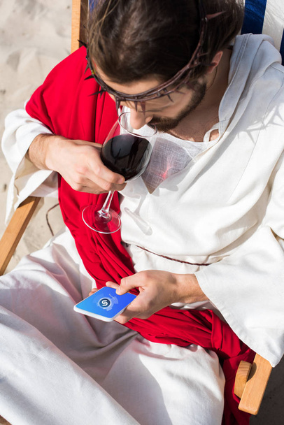 high angle view of Jesus resting on sun lounger, drinking wine and using smartphone with Shazam appliance in desert - Photo, image