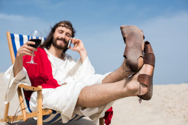 hymyilevä Jeesus lepää aurinkotuolissa viinilasillisen kanssa ja puhuu älypuhelimella aavikolla
 - Valokuva, kuva