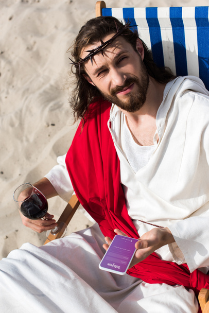 Jesus resting on sun lounger with glass of wine and holding smartphone with instagram appliance in desert - Фото, изображение