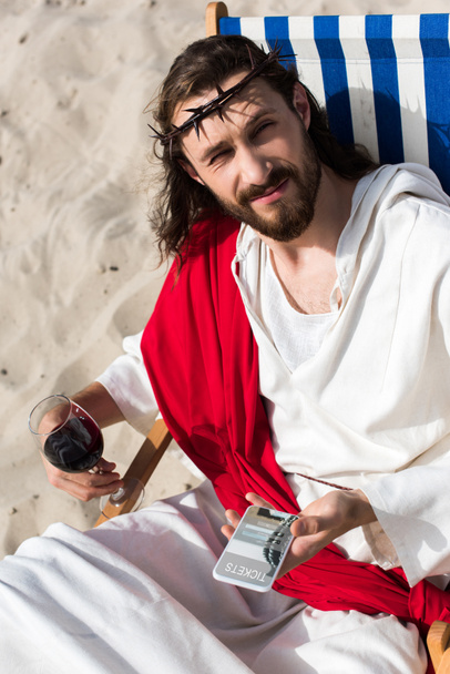 Jesus resting on sun lounger with glass of wine and holding smartphone with tickets website in desert - Foto, afbeelding