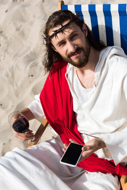 Jesus ruht sich auf Sonnenliege mit Glas Wein aus und hält Smartphone mit leerem Bildschirm in der Wüste - Foto, Bild