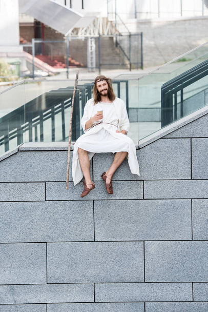 fröhlicher Jesus in Robe und Dornenkrone sitzt auf der Treppenseite und hält Kaffee to go - Foto, Bild
