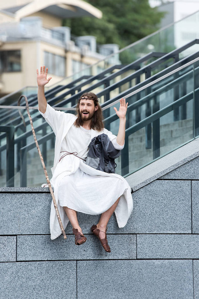 happy Jesus in robe and crown of thorns sitting on staircase side and waving hands - Foto, imagen