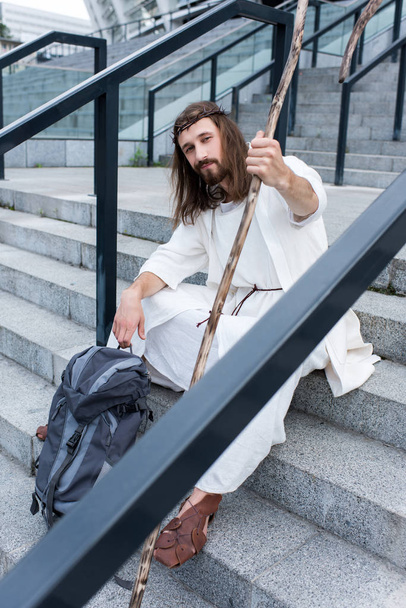 Jesus in robe and crown of thorns sitting on stairs with travel bag and staff, looking at camera - 写真・画像