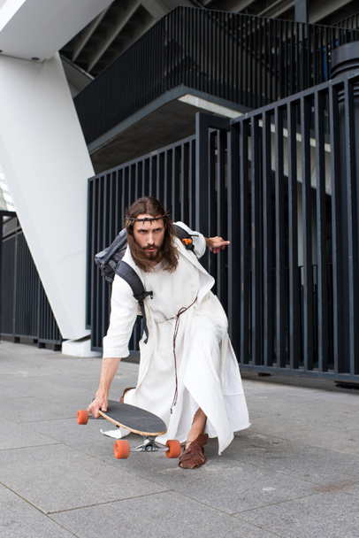 serious Jesus in robe and crown of thorns skating on longboard on street - Фото, изображение