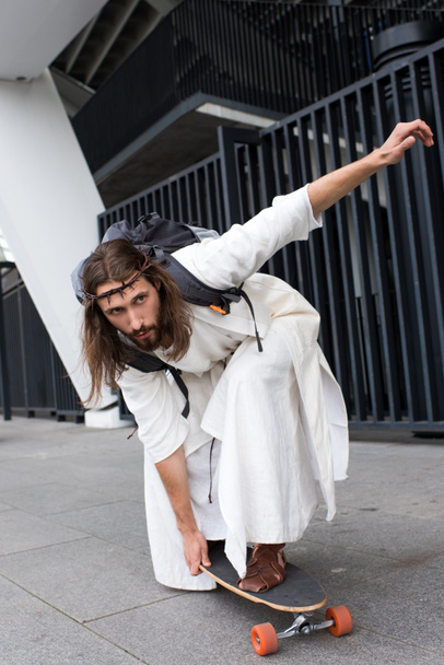 handsome Jesus in robe and crown of thorns skating on longboard on street - Fotografie, Obrázek
