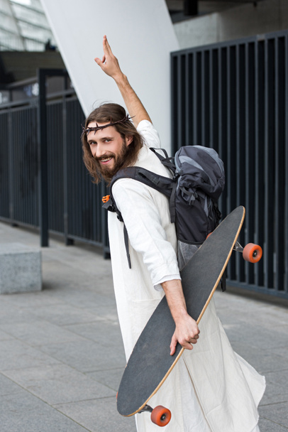 smiling Jesus in robe and crown of thorns walking with longboard and showing two fingers up - Фото, изображение
