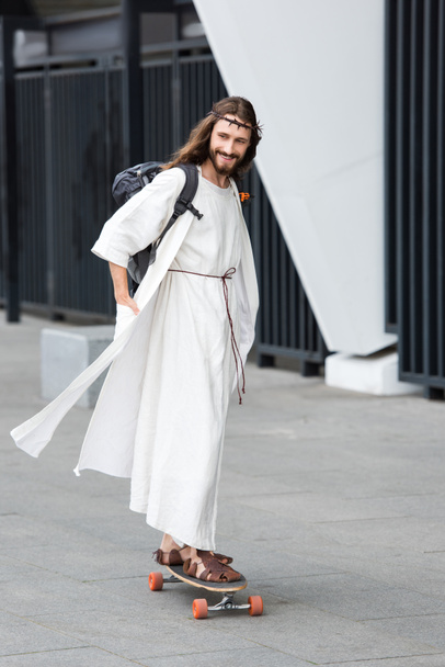 smiling Jesus in robe and crown of thorns skating on longboard on street  - Фото, изображение
