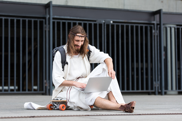 sorridente Gesù in veste e corona di spine seduto sullo skateboard e utilizzando il computer portatile in città
 - Foto, immagini