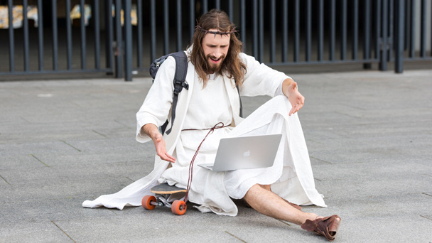 irritated Jesus sitting on skateboard and gesturing to laptop on street - Photo, Image