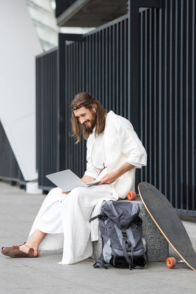 cheerful Jesus in robe and crown of thorns sitting on stone and using laptop on street - Zdjęcie, obraz