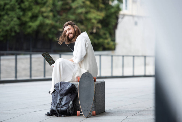 Jesus in robe and crown of thorns sitting on stone and holding laptop with html code on street - Zdjęcie, obraz