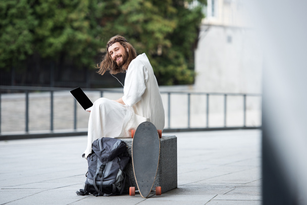 smiling Jesus in robe and crown of thorns sitting on stone and using laptop with blank screen on street - Foto, immagini