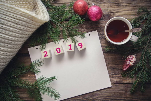 Beautiful Christmas card with new year 2019, tree branches, balls, scarf, cup of tea on a wooden rustic background, with copy space, top view - Photo, image