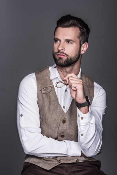 portrait of handsome bearded man holding glasses and posing in waistcoat, isolated on grey  - Fotó, kép