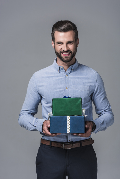 smiling stylish businessman holding gift boxes, isolated on grey - Fotoğraf, Görsel