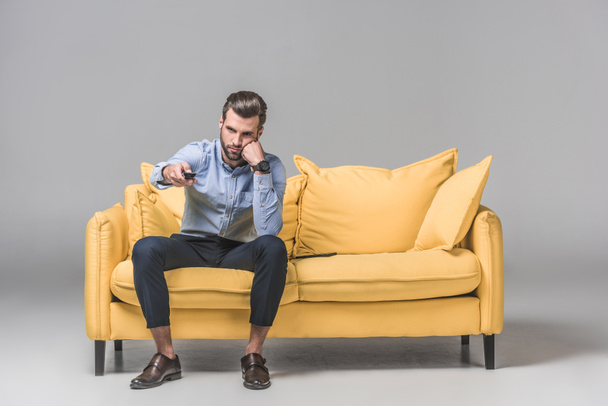 gelangweilte Mann mit Fernbedienung fernsehen und sitzen auf gelbem Sofa auf grau - Foto, Bild