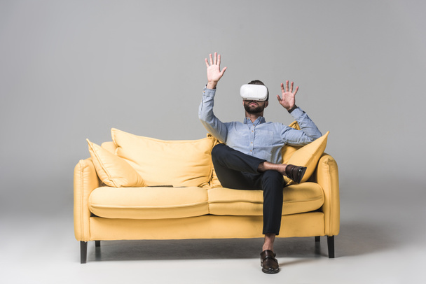 parrakas mies elehtii ja käyttää virtuaalitodellisuus kuulokkeet ja istuu keltainen sohva harmaa
 - Valokuva, kuva