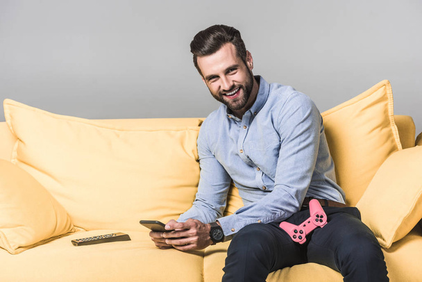 smiling man using smartphone and sitting on yellow sofa with joystick and remote control on grey - Photo, image