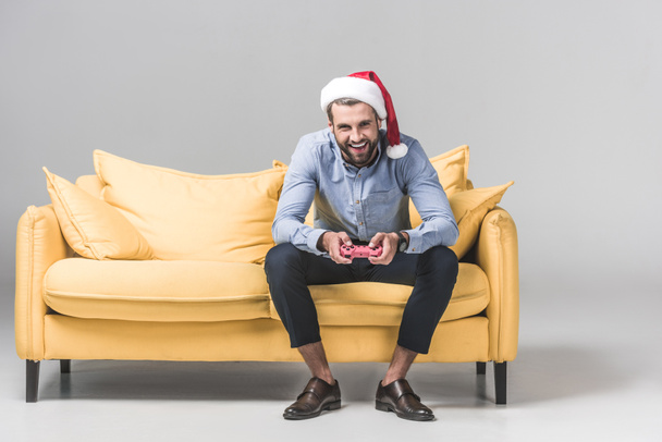 ευτυχισμένος άνθρωπος με το καπέλο santa παιχνίδι βίντεο με joystick ενώ κάθεστε στον καναπέ, σε γκρι - Φωτογραφία, εικόνα