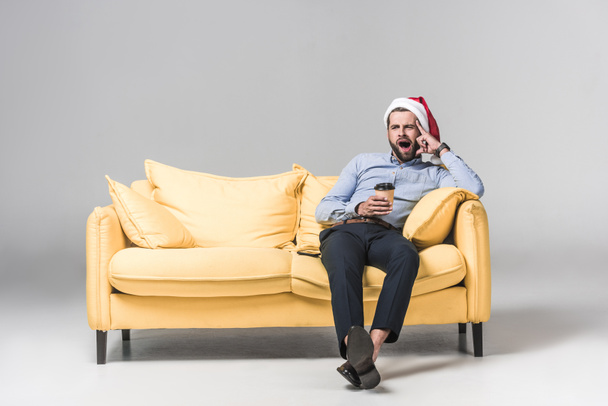hombre cansado en el bostezo del sombrero de santa, sosteniendo el café para ir y sentado en sofá amarillo en gris
 - Foto, Imagen