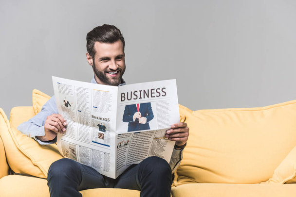 smiling bearded man reading business newspaper while sitting on sofa, on grey - Foto, imagen