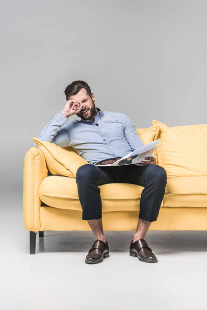 confused businessman with newspaper sitting on yellow sofa, on grey - Fotoğraf, Görsel
