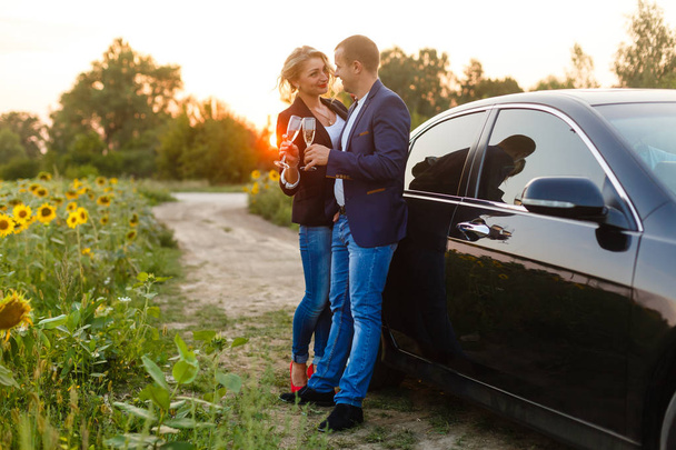 Young couple celebrating with champagne in sunflower field by car at sunset - Photo, image