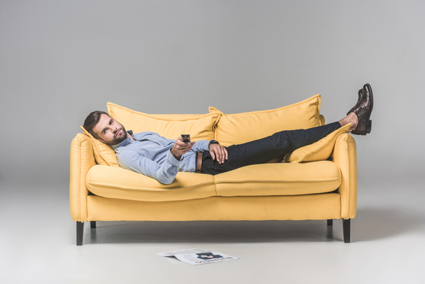 bearded man with remote control watching TV and resting on sofa with newspaper on floor, on grey - Fotografie, Obrázek