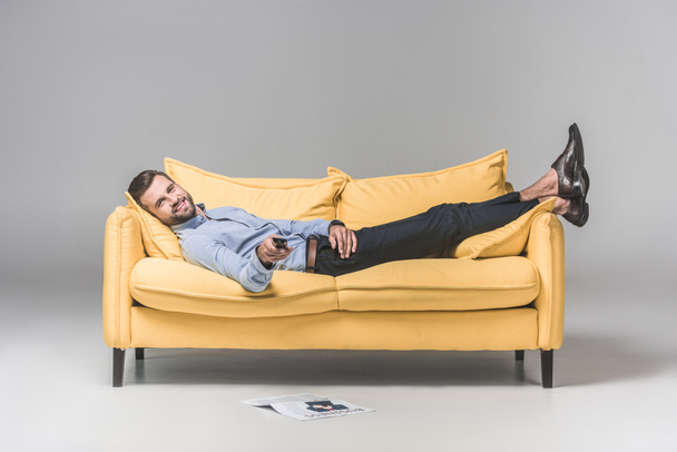 hombre sonriente con mando a distancia viendo la televisión y acostado en el sofá con periódico en el suelo, en gris
 - Foto, Imagen