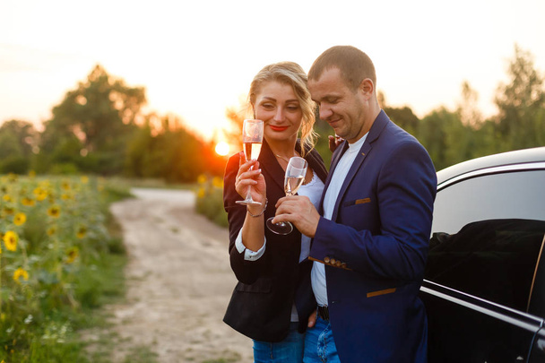Young couple celebrating with champagne in sunflower field by car at sunset - Foto, afbeelding