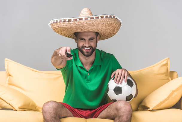Χαμογελαστός άνθρωπος σε μεξικάνικο σομπρέρο παρακολουθούν ποδόσφαιρο ταιριάζουν ενώ κάθεται σε κίτρινο καναπέ με μπάλα και απομακρυσμένο έλεγχο σε γκρι   - Φωτογραφία, εικόνα