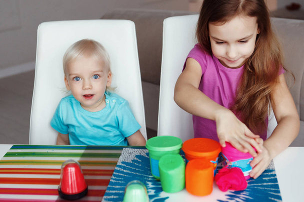 İki mutlu çocuk sarışın erkek ve kız hamuru ile ve evde masada oturan pembe, kırmızı ve yeşil su bardağı ile oynama - Fotoğraf, Görsel