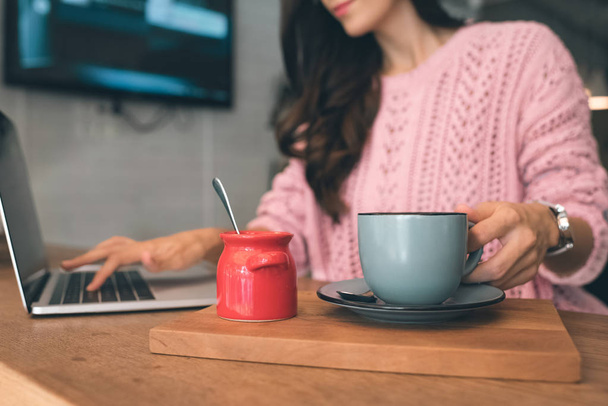 bijgesneden afbeelding van vrouwelijke freelancer zitten met laptop en koffie kopje aan tafel in café - Foto, afbeelding