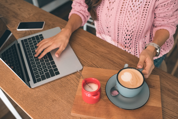 imagen recortada del freelancer femenino que trabaja en el ordenador portátil mientras está sentado en la mesa con teléfono inteligente y taza de café en la cafetería
  - Foto, Imagen