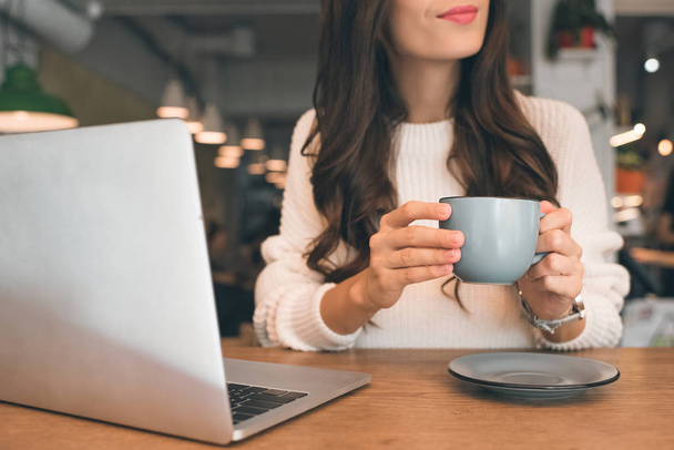 частичный вид женщины-фрилансера, сидящей с ноутбуком и чашкой кофе за столом в кафе
 - Фото, изображение