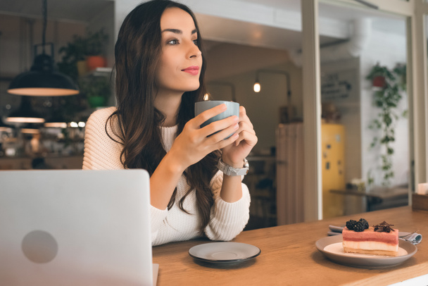 aantrekkelijke vrouwelijke freelancer zitten met laptop, taart en koffie kopje aan tafel in café - Foto, afbeelding