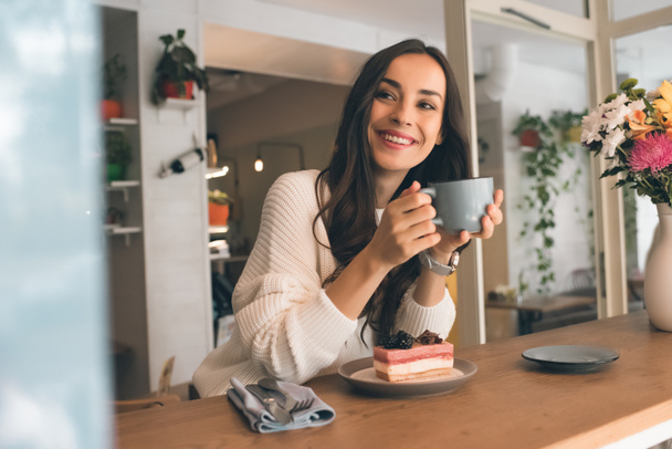 Szczęśliwa młoda kobieta z filiżanki kawy, siedzi przy stole z sernik w kawiarni - Zdjęcie, obraz