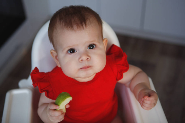 Şirin bir bebek tombul yanakları kırmızı elbiseli bir beyaz çocuk sandalyeye oturur, bir yeşil elma yiyor ve kameraya baktı. Yakın çekim. - Fotoğraf, Görsel