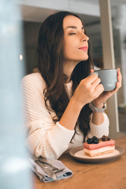 приваблива молода жінка з закритими очима тримає чашку кави і сидить за столом з чізкейком в кафе
 - Фото, зображення