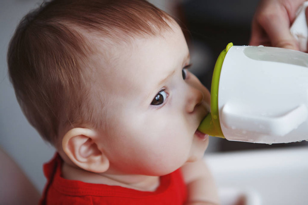 A cute baby in a red dress with puffy cheeks sits on a white children's chair. Drinks water from the children's cup. Close-up. - Foto, afbeelding