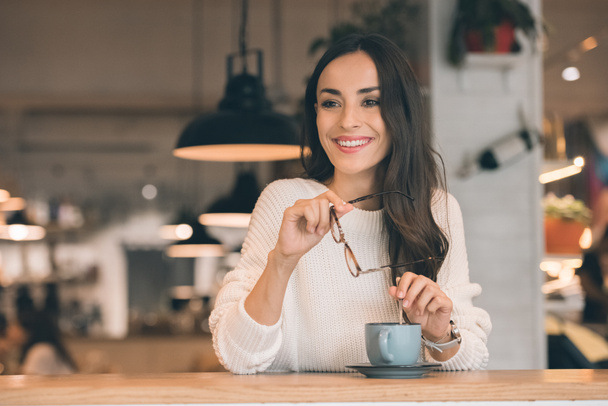 kafede kahve fincanı ile masada otururken gözlük tutan gülümseyen genç kadın - Fotoğraf, Görsel