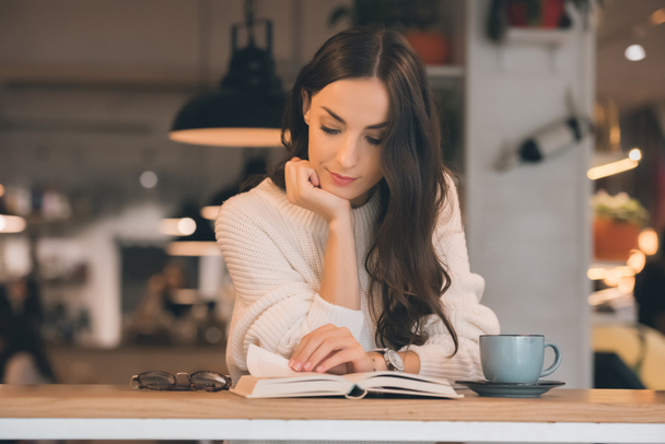 Konzentrierte junge Frau liest Buch am Tisch mit Kaffeetasse im Café - Foto, Bild