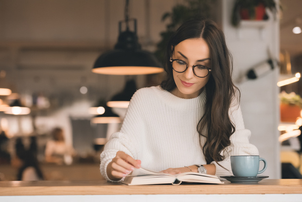 attrayant jeune femme dans les lunettes de lecture livre à table avec tasse de café dans le café
 - Photo, image
