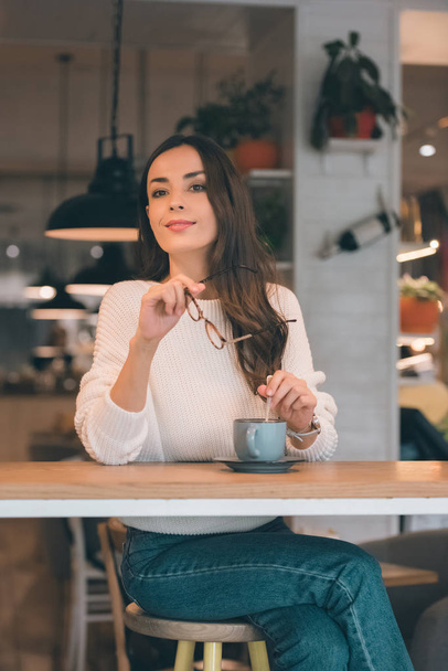 beautiful young woman looking away and holding eyeglasses while sitting at table with coffee cup in cafe - Foto, afbeelding